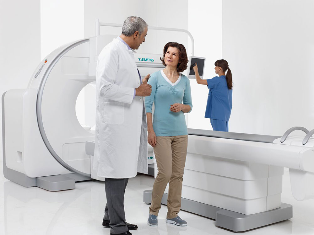 Patient standing next to a Siemens CT scanner while a doctor discusses the procedure with her.