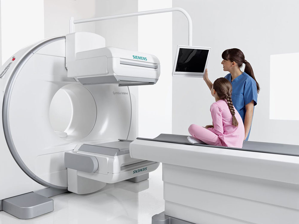 A Siemens MRI machine with a patient lying on the scanner table while a clinician and a girl in pink await the procedure, the technician pointing at a wall monitor to review imaging.